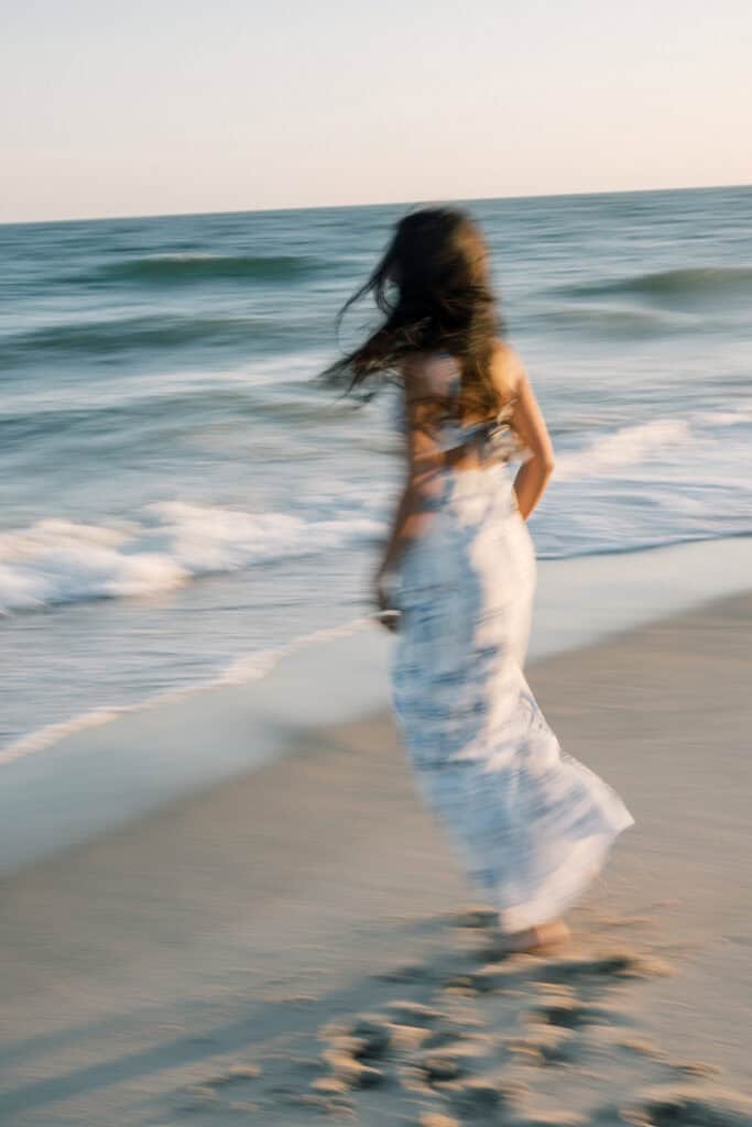 A woman in a white dress walks along the shoreline, with motion blur, as waves approach the sandy beach under a clear sky—captured by photographer Kaity Farrell for Nantucket senior portraits.
