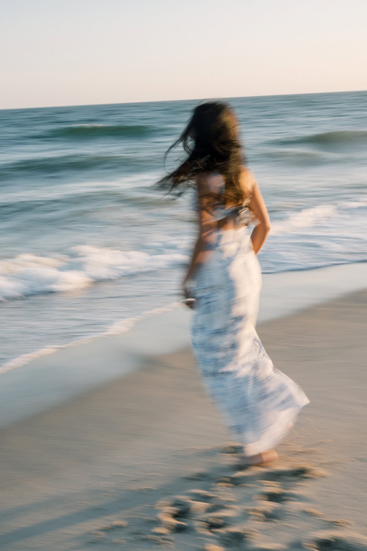 A woman in a white dress walks along the shoreline, with motion blur, as waves approach the sandy beach under a clear sky—captured by photographer Kaity Farrell for Nantucket senior portraits.