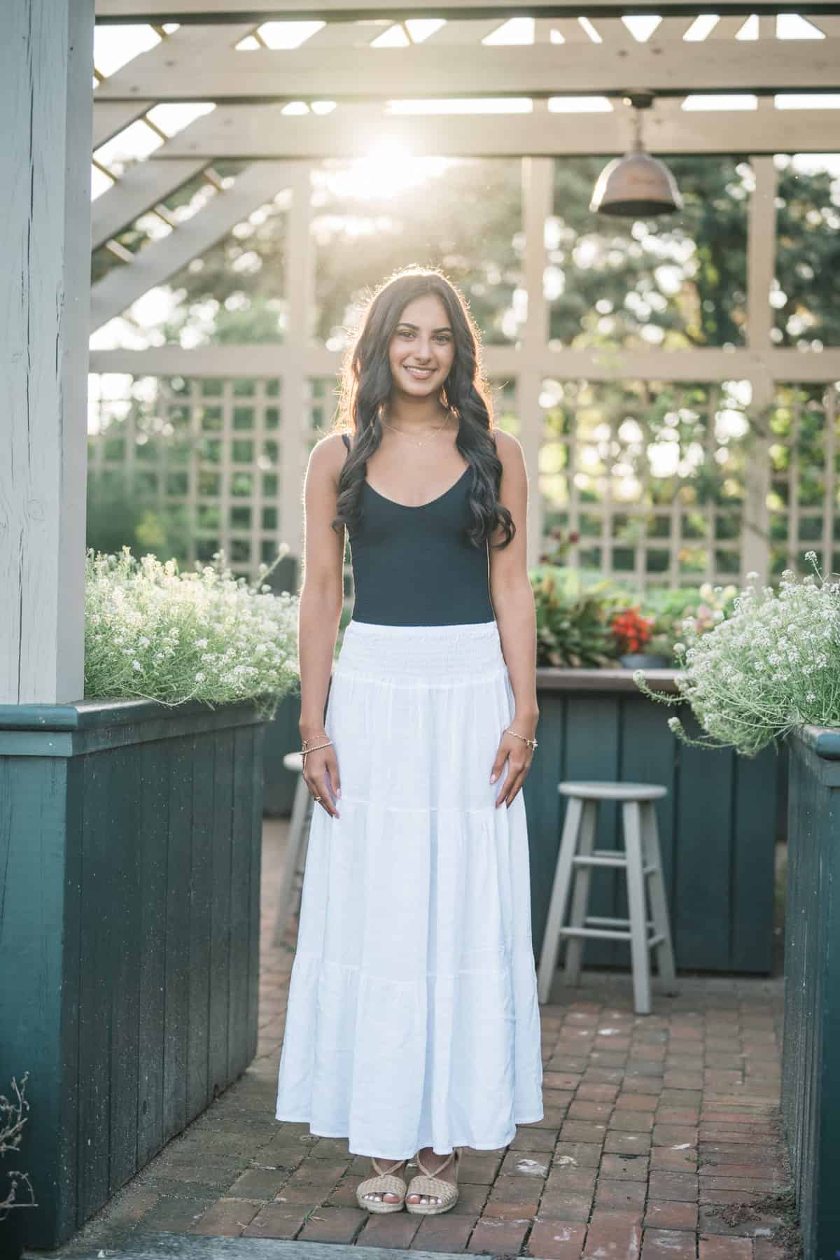 A young woman standing outdoors on a brick patio in a black sleeveless top and long white skirt, with plants and sunlight in the background—captured during a high school senior photo session by Nantucket photographer Kaity Farrell.