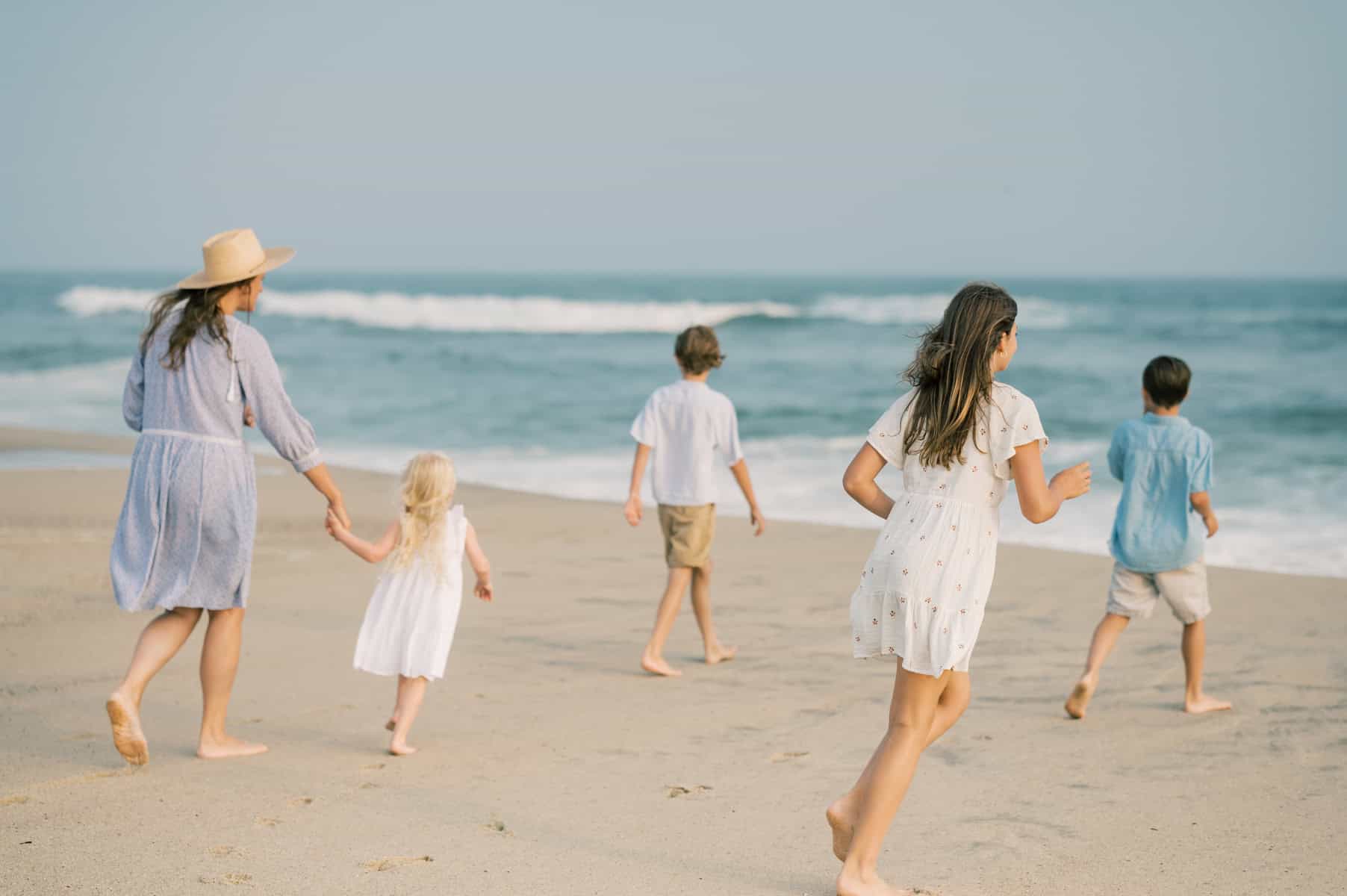 A mother and four children walk and run barefoot along a sandy beach near the ocean on Nantucket, with waves in the background—captured by Nantucket Family Portrait Photographer Kaity Farrell.