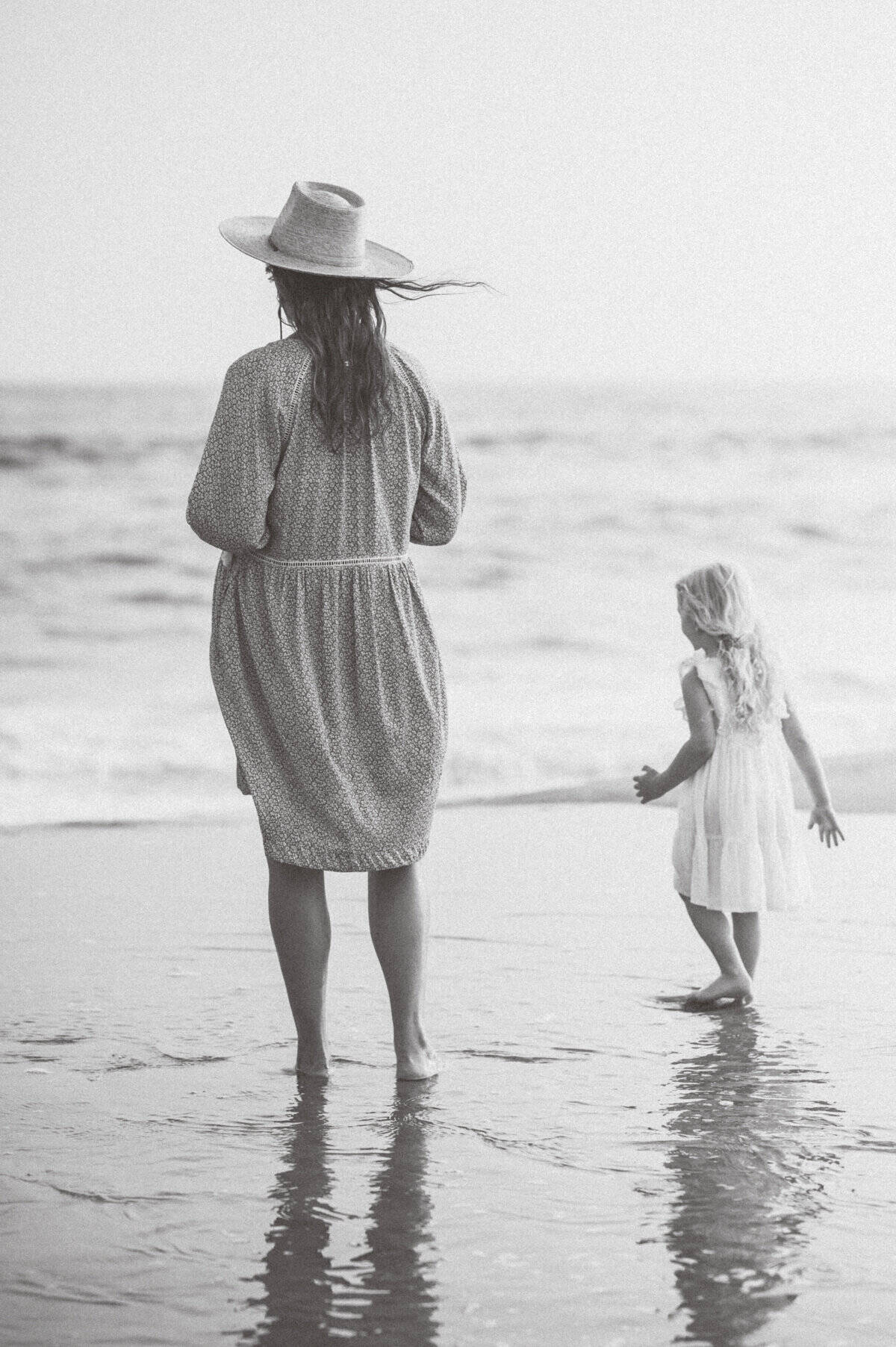 A woman in a dress and hat stands on a beach next to a young girl walking toward the water; both have their backs to the camera, capturing a timeless moment from a Nantucket family photo session by photographer Kaity Farrell.