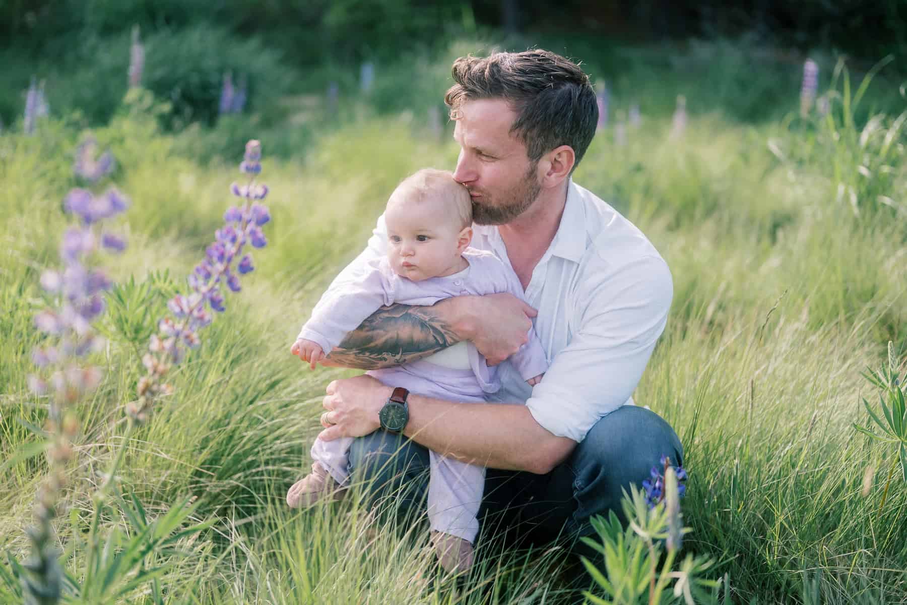 A father kneels in a grassy field, holding a baby in his arms and kissing the baby's head as purple wildflowers bloom nearby—a tender moment captured during a family portrait session by Nantucket photographer Kaity Farrell.
