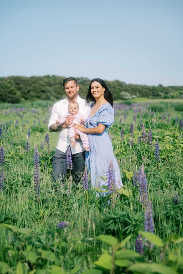 A mother and father stand in a green field with purple lupine flowers, holding a baby. Captured by photographer Kaity Farrell, this Nantucket family photo beautifully frames trees and sky in the background.