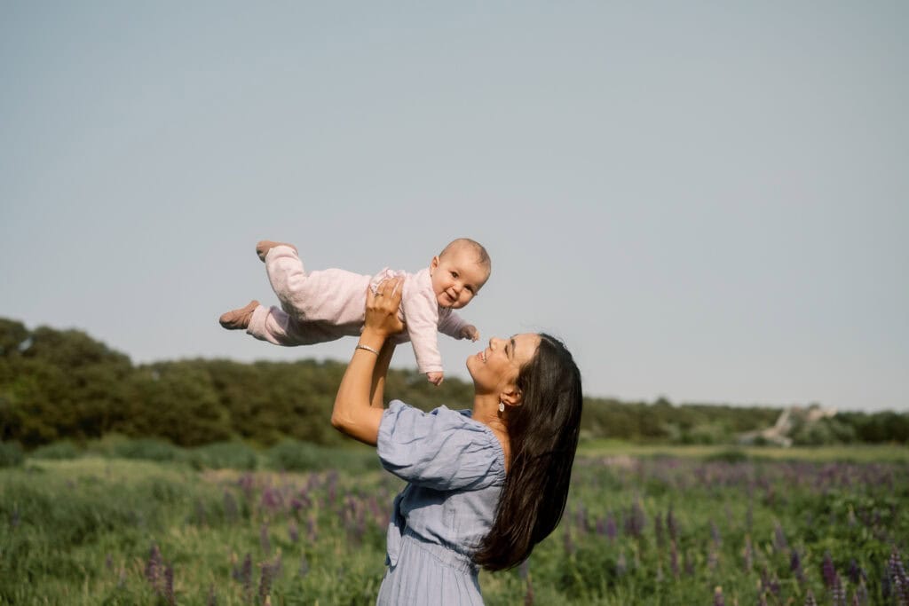 A woman in a blue dress lifts a smiling baby above her head in an outdoor Nantucket flower field, with green grass and trees in the background—captured beautifully by photographer Kaity Farrell.