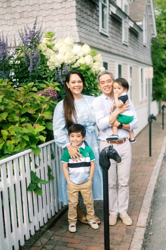A family of four stands on a brick sidewalk by a white fence and greenery, with a house in the background. The adults smile as they pose with two young children.