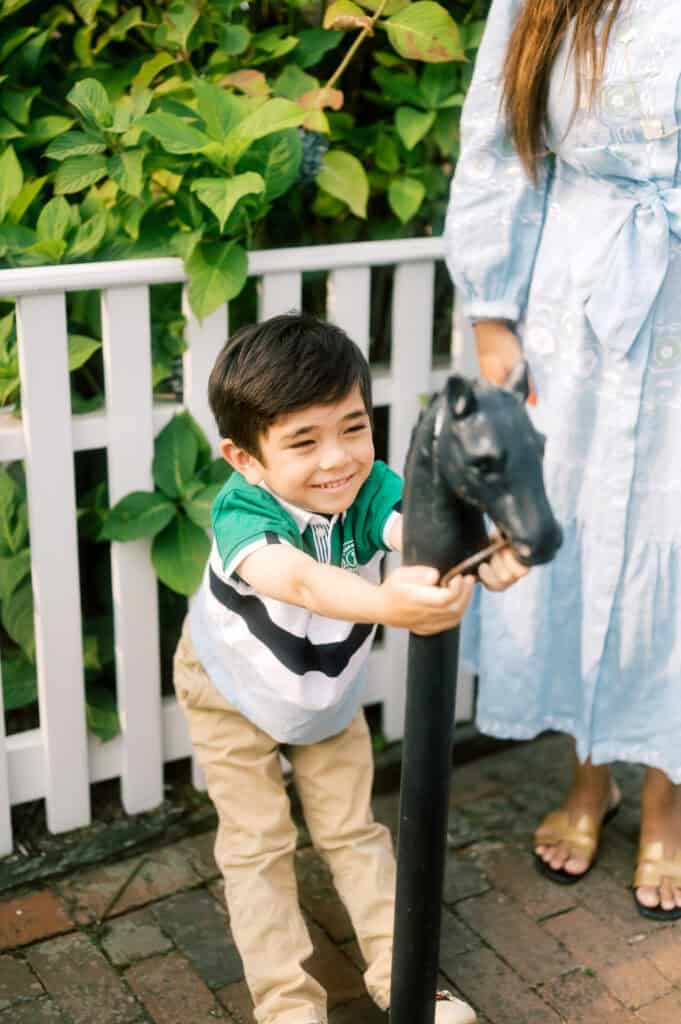 A young boy smiles while holding onto a black horse head post, standing next to an adult in a light blue dress by a white fence and green plants during a Nantucket family portrait session with photographer Kaity Farrell.