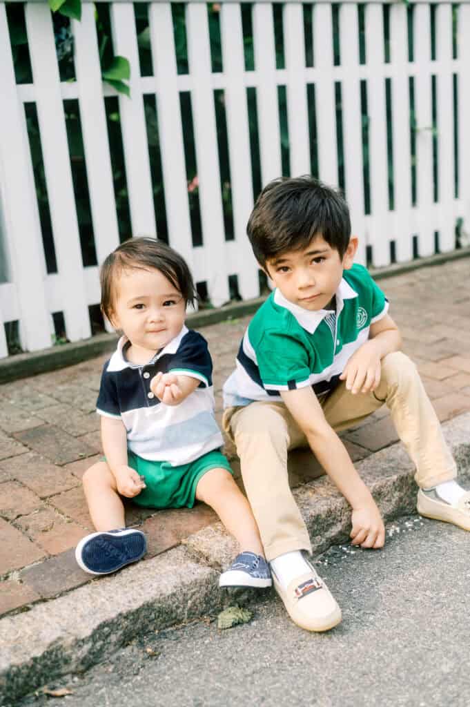 Two young boys sit on a curb in front of a white picket fence. Captured during a Nantucket family portrait session by photographer Kaity Farrell, the barefoot younger boy smiles while his older brother sits with one knee up, gazing at the camera.