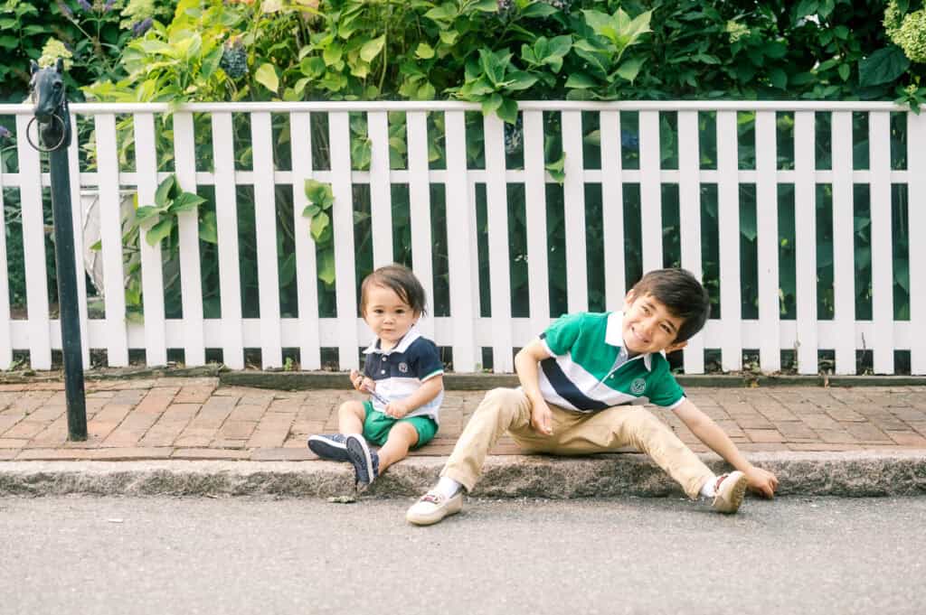 Two young children sit on a curb in front of a white picket fence; one sits upright while the other leans back with a playful expression, captured during a Nantucket family portrait session by photographer Kaity Farrell.