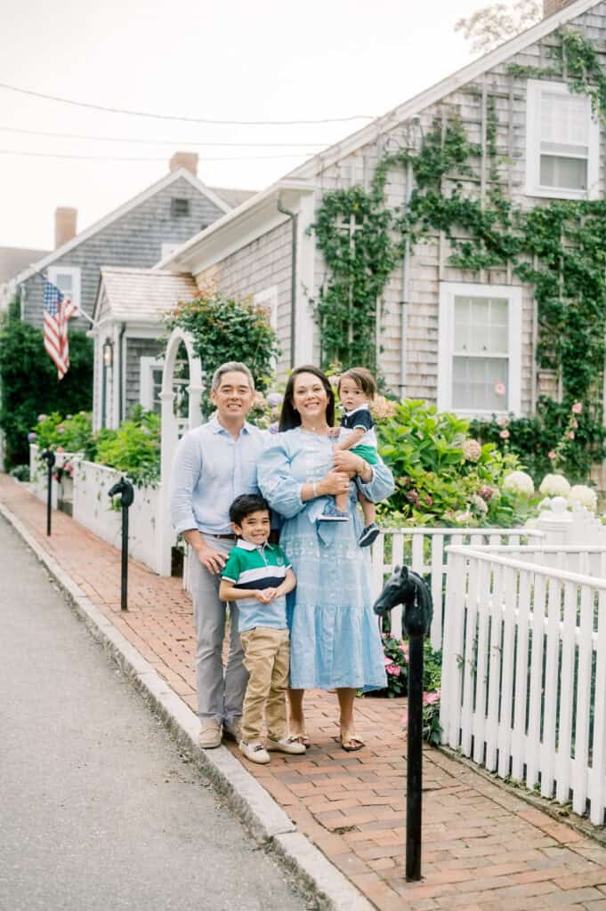 A family of four poses on a brick sidewalk in front of a white picket fence and a vine-covered house with an American flag during their Nantucket family photo session with photographer Kaity Farrell.