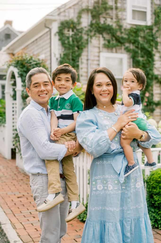 A family of four stands outside in front of a house with greenery, smiling at the camera during a family photo session with Nantucket photographer Kaity Farrell. The parents each hold a young child.