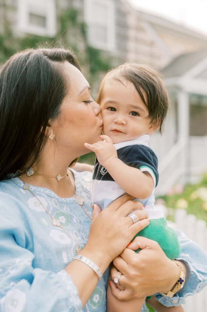 A woman holds and kisses a baby on the cheek while standing outside in front of a house with greenery and a white picket fence during a Nantucket family portrait session with photographer Kaity Farrell.