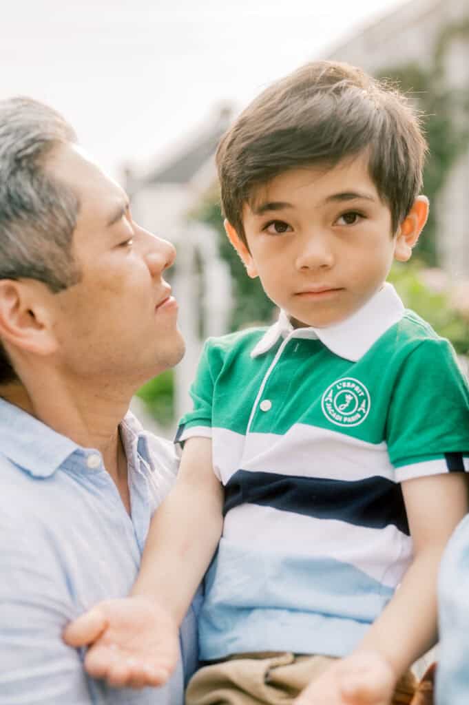 A father holds a young boy in a green, blue, and white striped polo shirt during a Nantucket family portrait session. Both are outdoors, with the child looking directly at the camera, expertly captured by photographer Kaity Farrell.