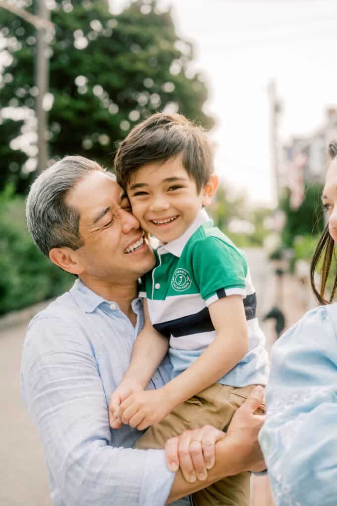 An older man smiles and hugs a young boy outdoors as he laughs in a green and white striped shirt, captured during a Nantucket family portrait session by photographer Kaity Farrell.