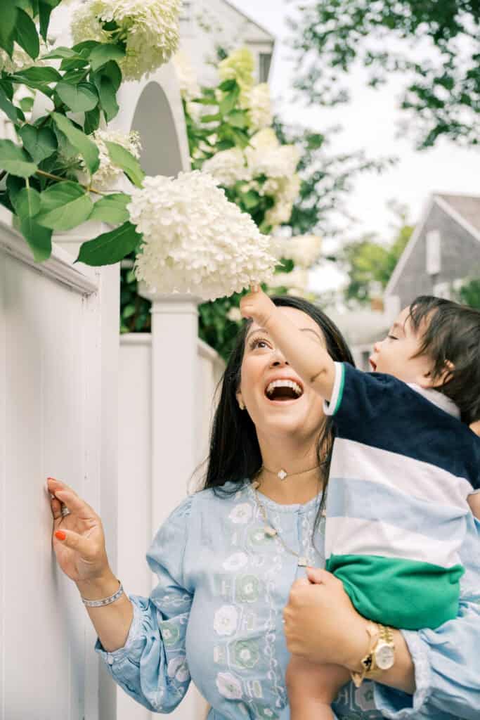 During a Nantucket family portrait session, a woman holds her young child as they both look up and reach toward white hydrangea flowers over a white fence outdoors, beautifully captured by photographer Kaity Farrell.