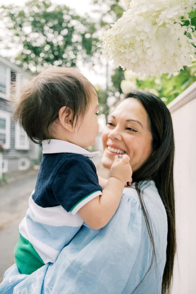 A woman smiles while holding a young child outdoors near white flowers, with houses and greenery in the background during a Nantucket family portrait session by photographer Kaity Farrell.