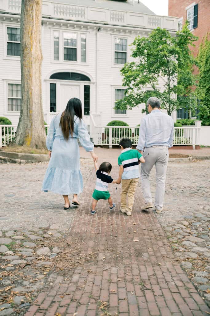 Two adults and two children walk hand in hand on a cobblestone path toward a white house, captured during a Nantucket family portrait session by photographer Kaity Farrell, with greenery surrounding the black door and white fence.