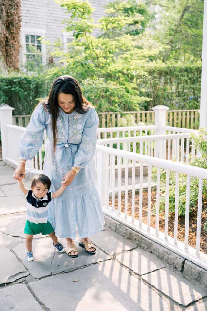 A woman helps a toddler walk on a stone path outside, surrounded by a white fence and greenery, during a Nantucket family portrait session with photographer Kaity Farrell.