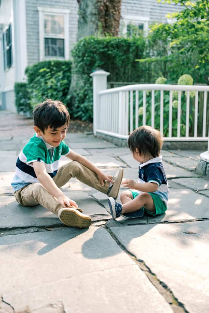 During a Nantucket family portrait session, two young children sit on a stone walkway outdoors, with the older child helping the younger one put on shoes. A white fence and greenery are in the background. Photographed by Kaity Farrell.