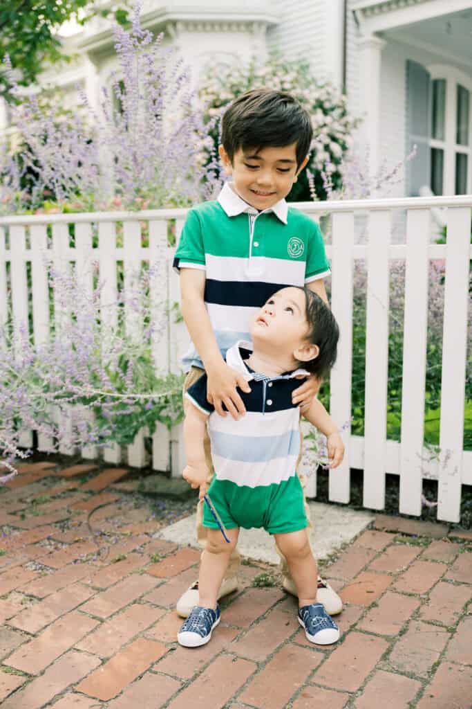 Two young boys stand together on a brick walkway in front of a white fence and house, with purple flowers nearby. Captured during a Nantucket family portrait session, the older boy hugs the younger, who looks up at him.