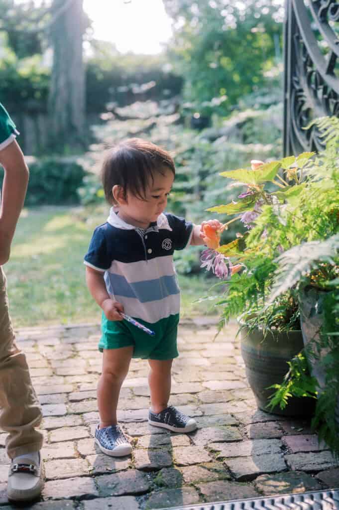 During a Nantucket family photo session, a young child stands on a stone path in a garden, reaching out to touch flowers on a plant while holding an object in their other hand, beautifully captured by photographer Kaity Farrell.