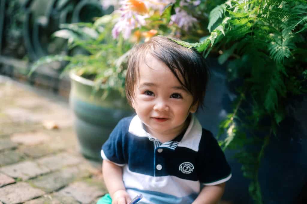 A young child with dark hair sits outdoors on a brick path, wearing a navy and white polo shirt, with green plants and flowers in the background—captured during a Nantucket family photo session by photographer Kaity Farrell.