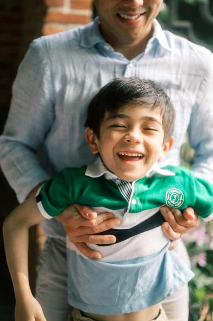 A man holds a smiling young boy around the waist outdoors during a Nantucket family photo session. The boy laughs in his green and white shirt as sunlight shines on them, captured perfectly by photographer Kaity Farrell.