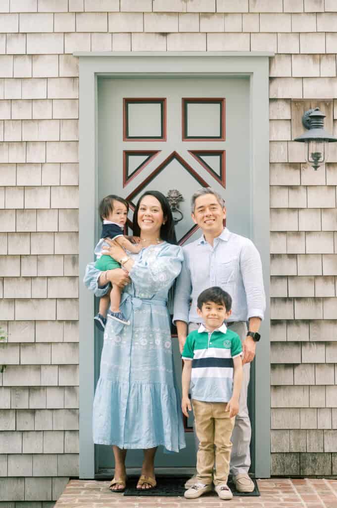 A family of four stands smiling in front of a house door; the mother holds a young child while the father and an older child stand beside them during their Nantucket family photo session with photographer Kaity Farrell.