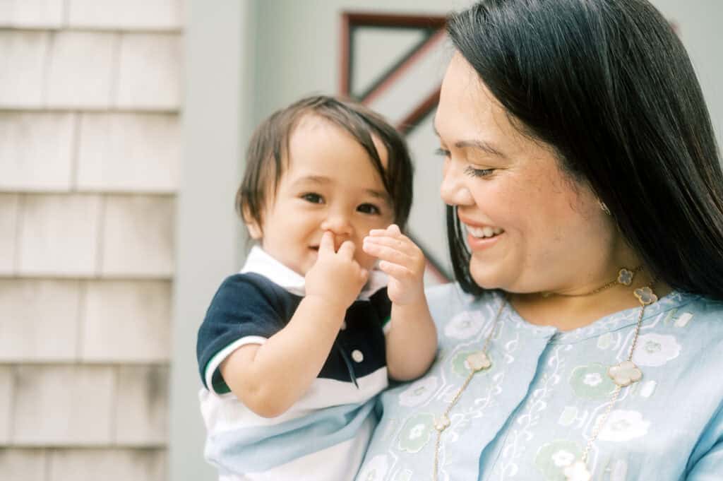 A woman smiles while holding a baby in a striped shirt who is touching their mouth during a Nantucket family photo session with photographer Kaity Farrell.
