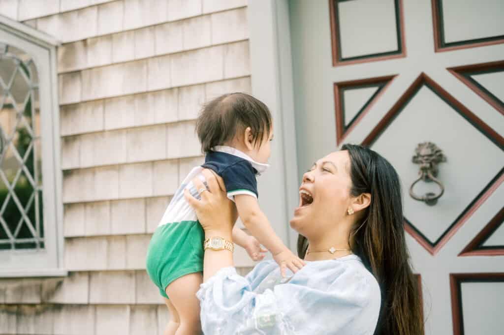 A woman stands outside a house, smiling as she lifts a baby in the air during a Nantucket family photo session with photographer Kaity Farrell.