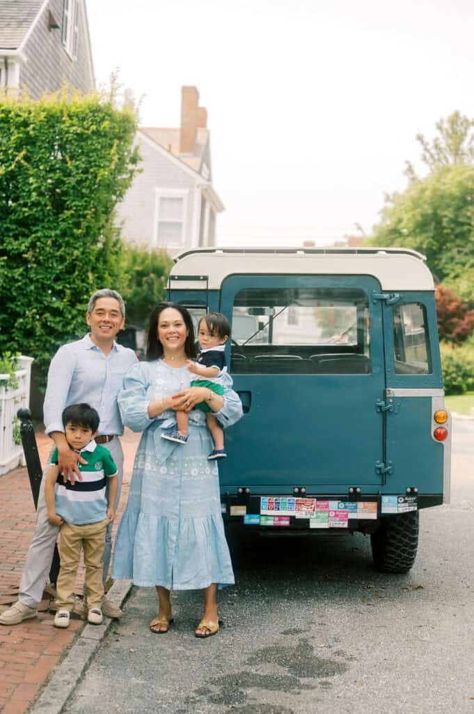 A family of four stands on a brick sidewalk next to a blue vehicle parked on a residential street, with houses and greenery in the background during their Nantucket family photo session with photographer Kaity Farrell.