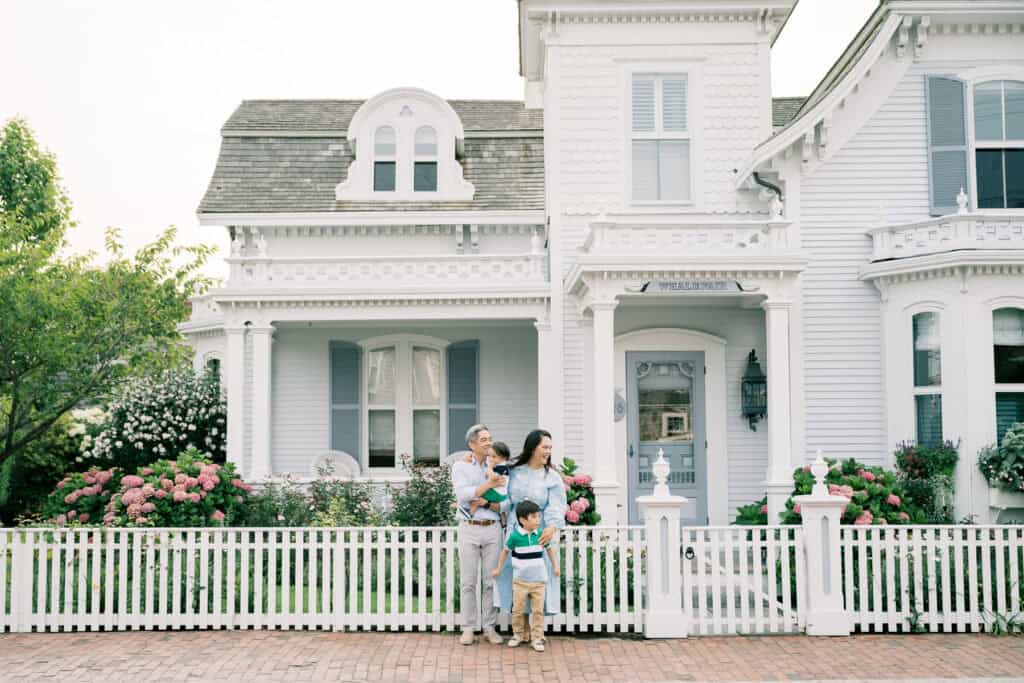 A family of four stands in front of a white Victorian-style house with a picket fence and blooming hydrangeas, captured during a family photo session by Nantucket photographer Kaity Farrell.