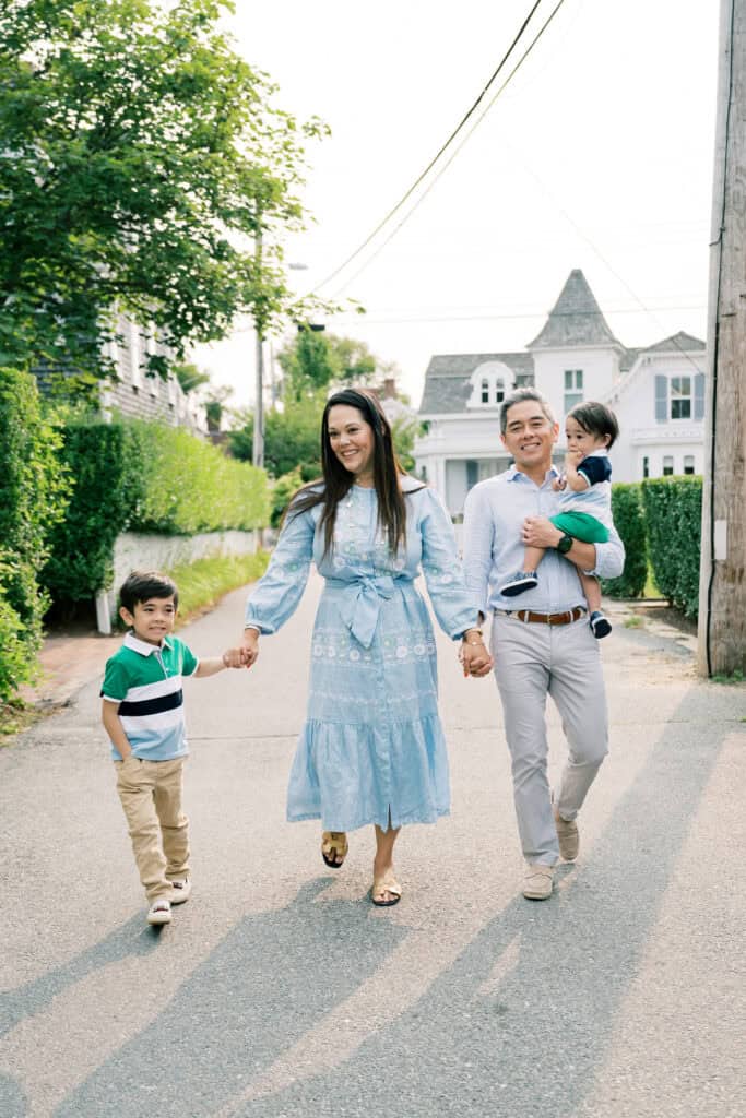 A family of four walks down a residential street, with the mother and father holding hands and each carrying one child, beautifully captured during a Nantucket family photo session by photographer Kaity Farrell.