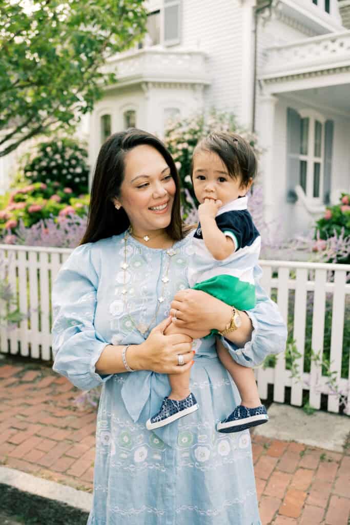 A woman in a blue dress holds a young child outside in front of a white picket fence and a house with flowers and greenery, captured during a family photo session by Nantucket photographer Kaity Farrell.