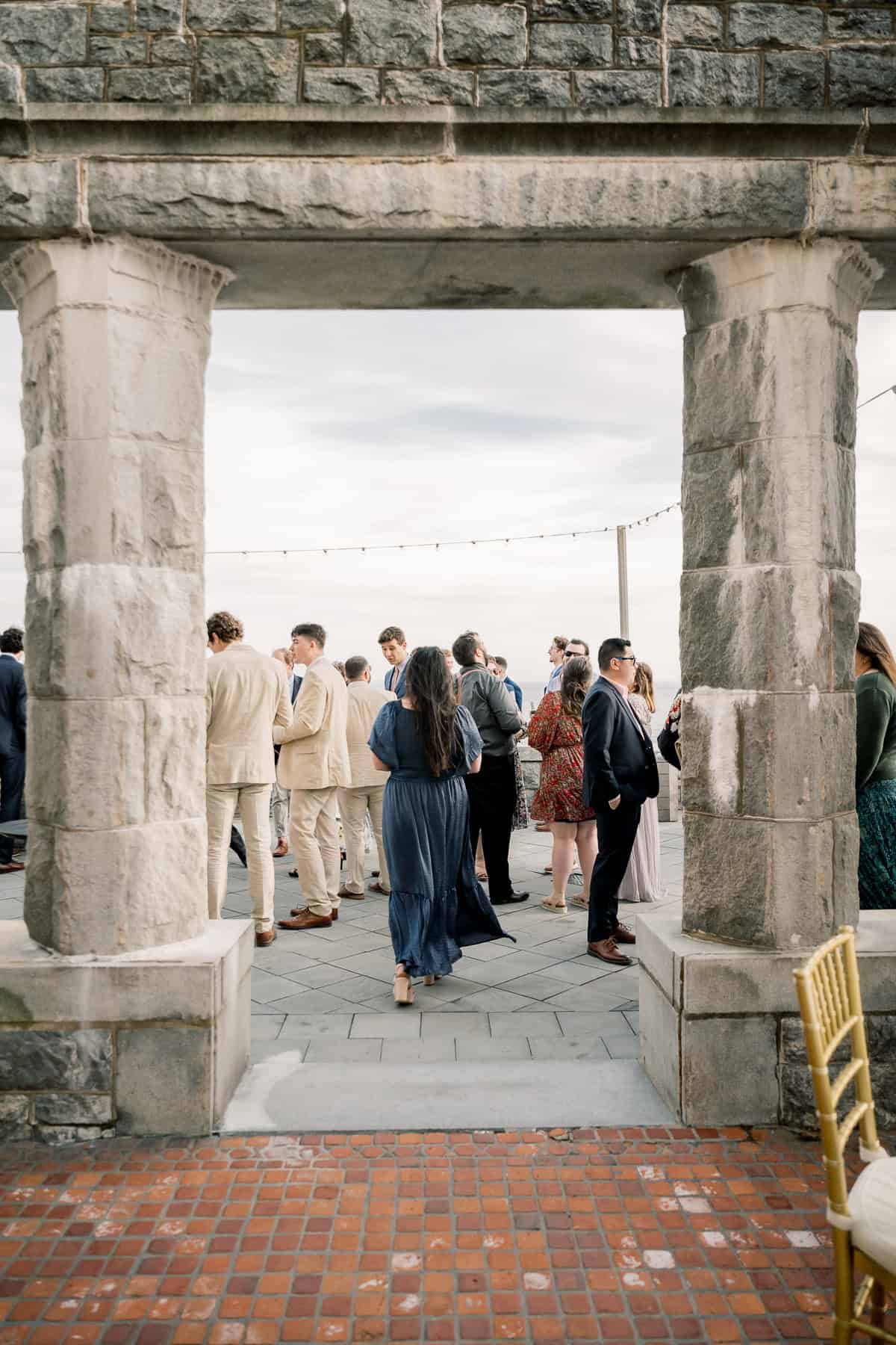 A group of people in formal attire stand and converse on an outdoor patio, viewed through a stone archway—an elegant moment from a New England wedding captured by photographer Kaity Farrell.