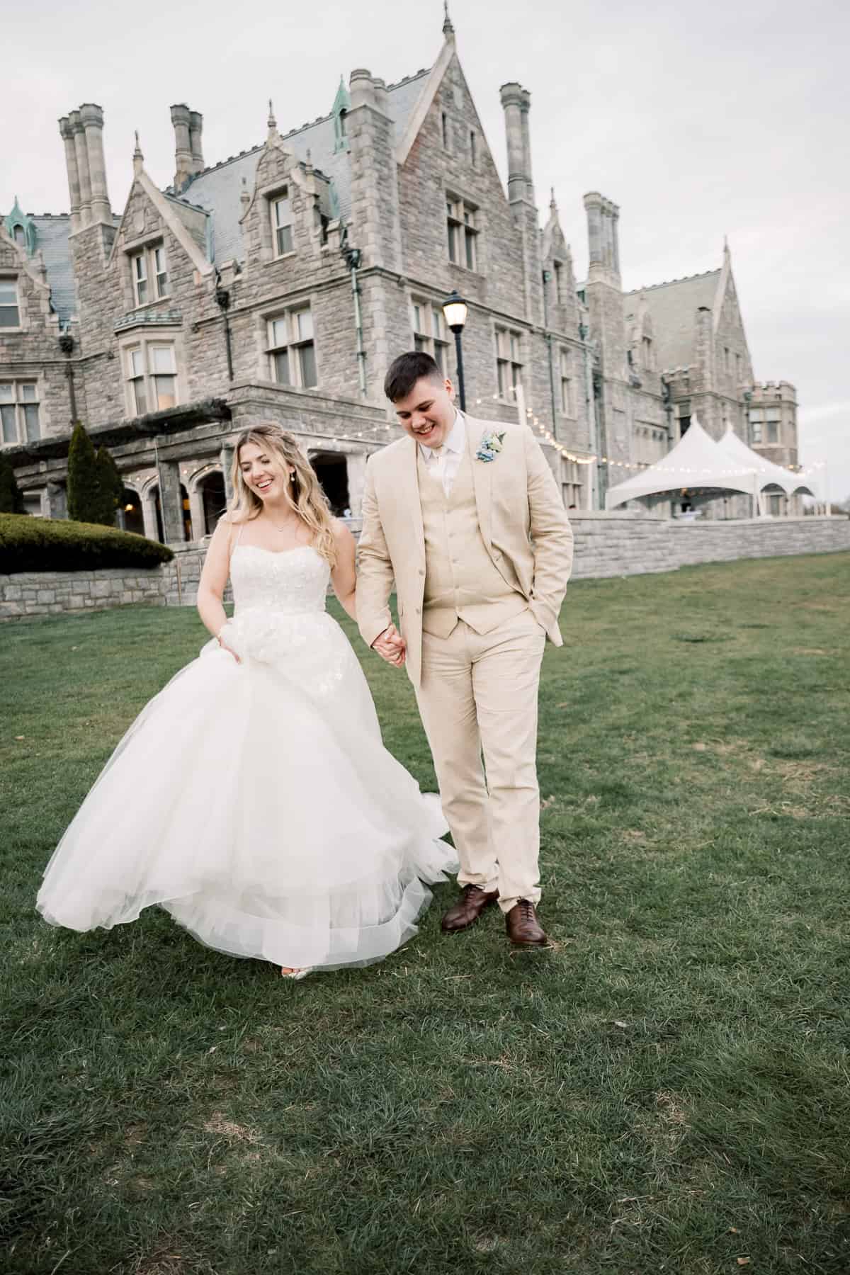 A bride and groom in wedding attire walk hand in hand on grass before a grand stone building with gothic architecture, beautifully captured by New England wedding photographer Kaity Farrell.