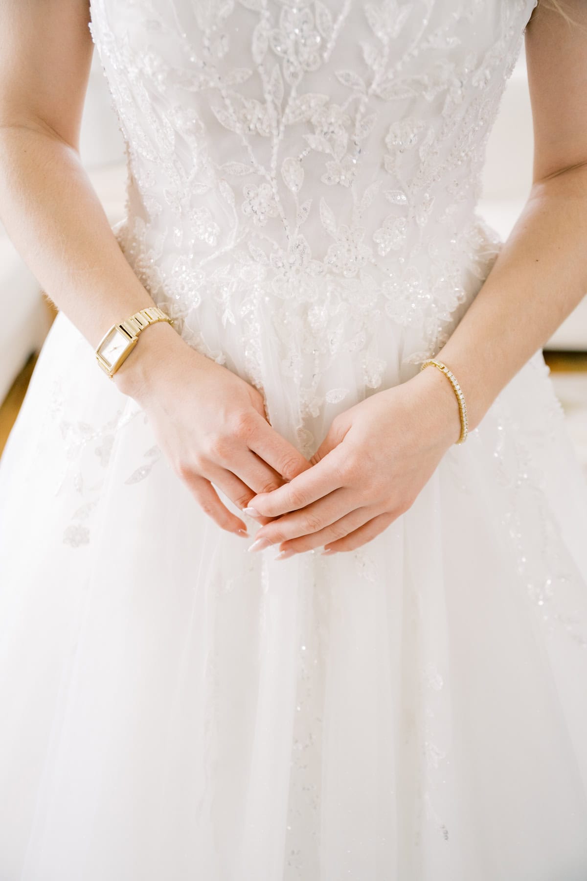 A bride wearing an ornate white wedding dress with lace details, a gold watch, and a bracelet stands with hands gently clasped—captured in elegant detail by photographer Kaity Farrell at a classic New England wedding.