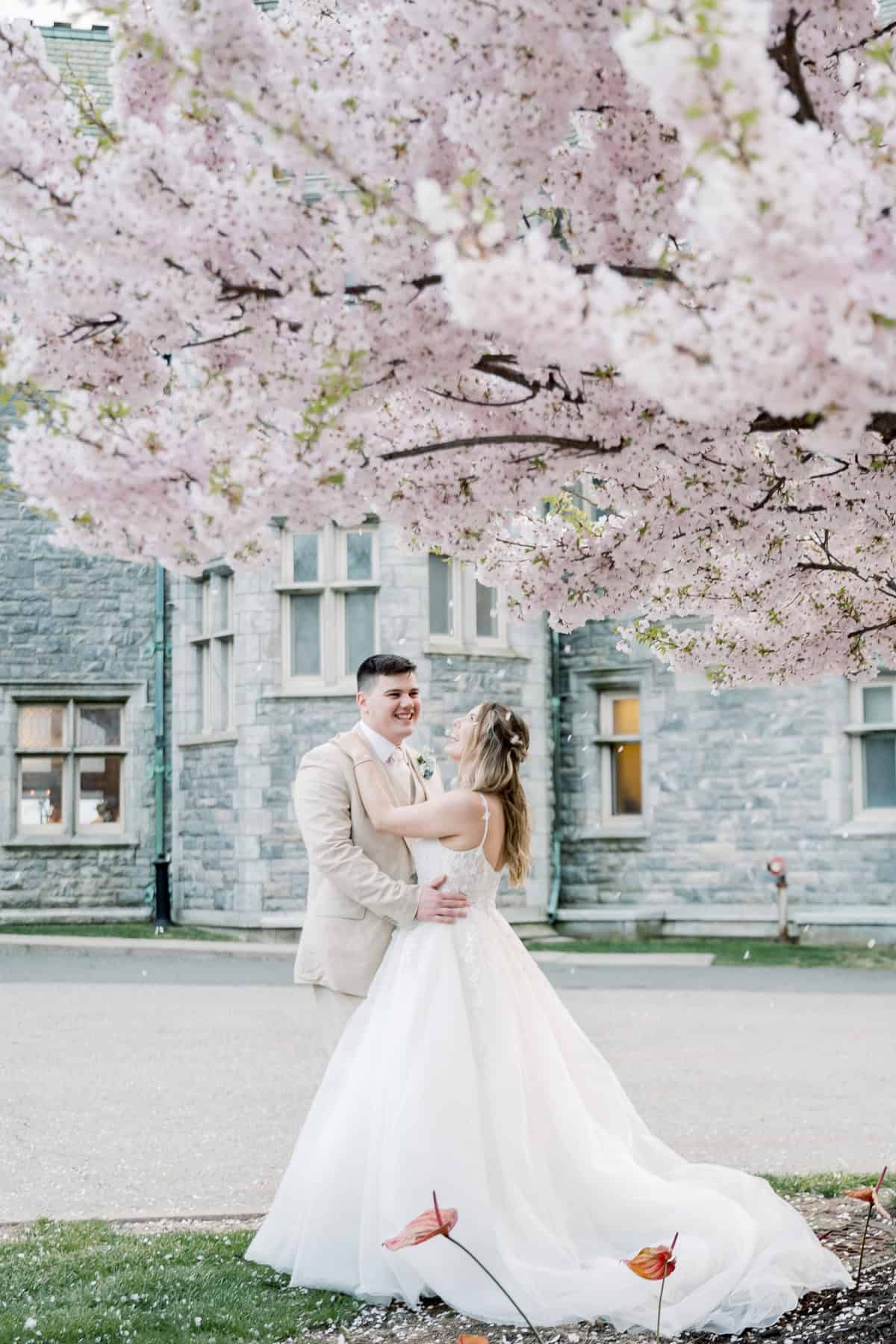 A bride and groom stand beneath a blooming cherry blossom tree, smiling at each other in front of a stone building at their New England wedding, beautifully captured by photographer Kaity Farrell.