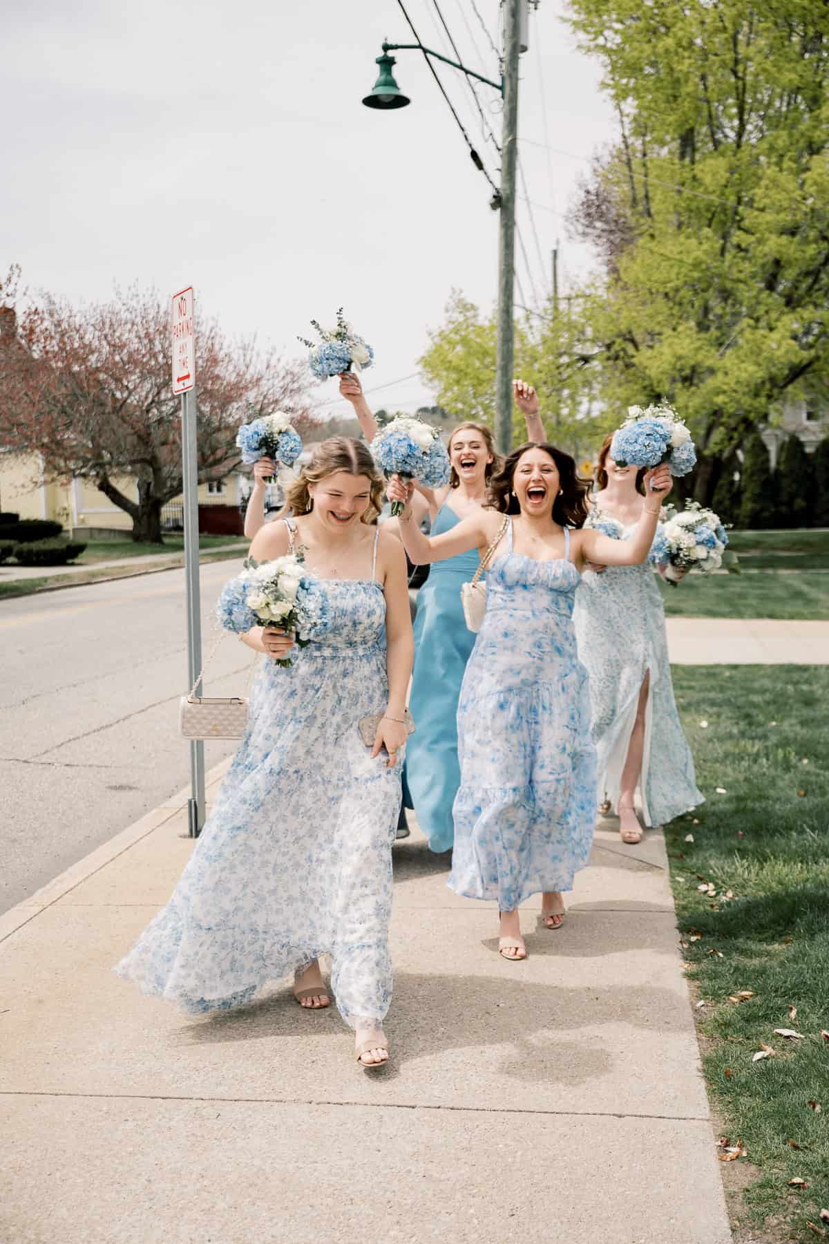 Four women in blue floral dresses holding bouquets walk and smile on a sunny, tree-lined New England wedding day, beautifully captured by photographer Kaity Farrell.