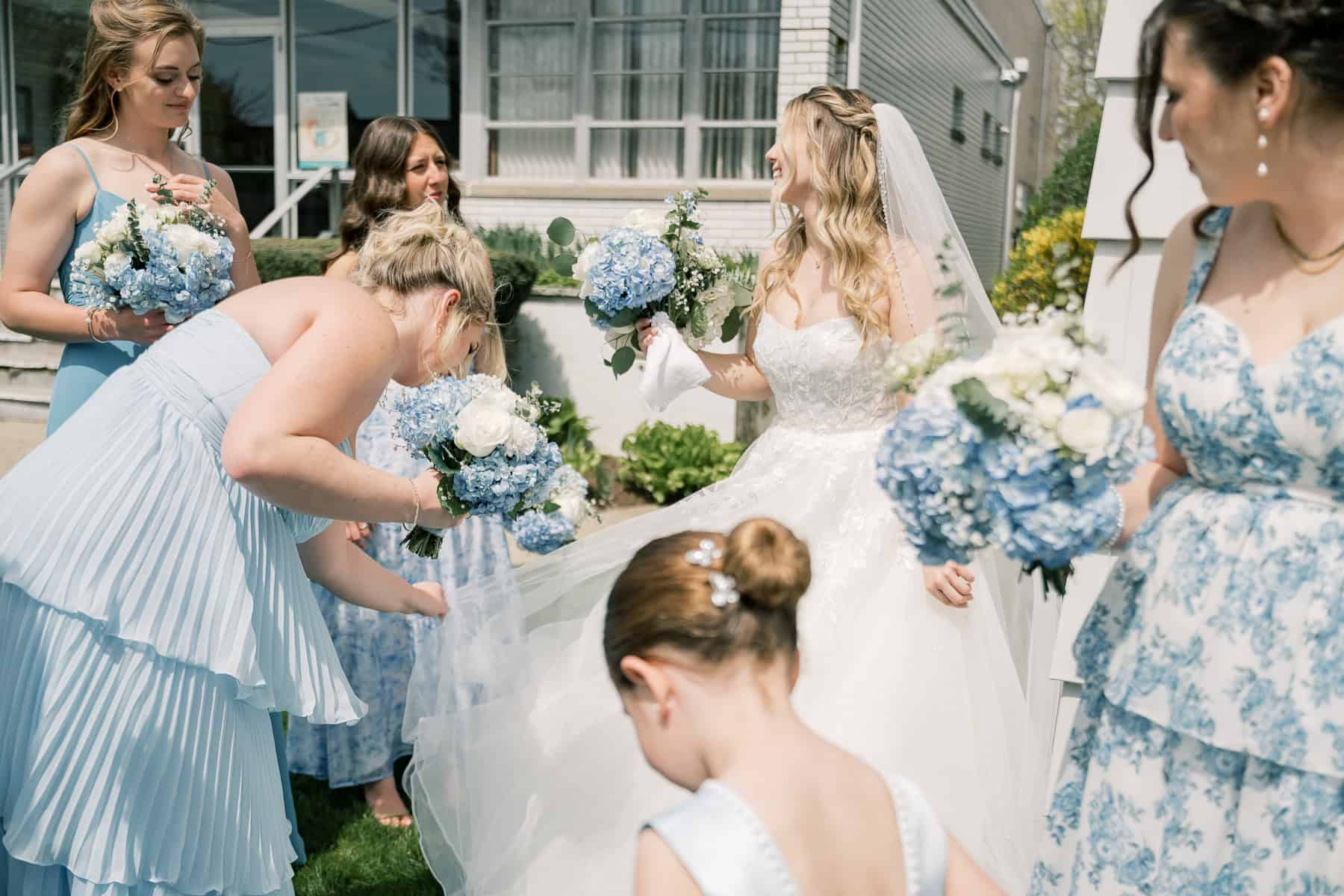 Captured by photographer Kaity Farrell at a charming New England wedding, a bride in white stands outdoors with her bridesmaids and flower girl in blue dresses, all holding bouquets of blue and white flowers.