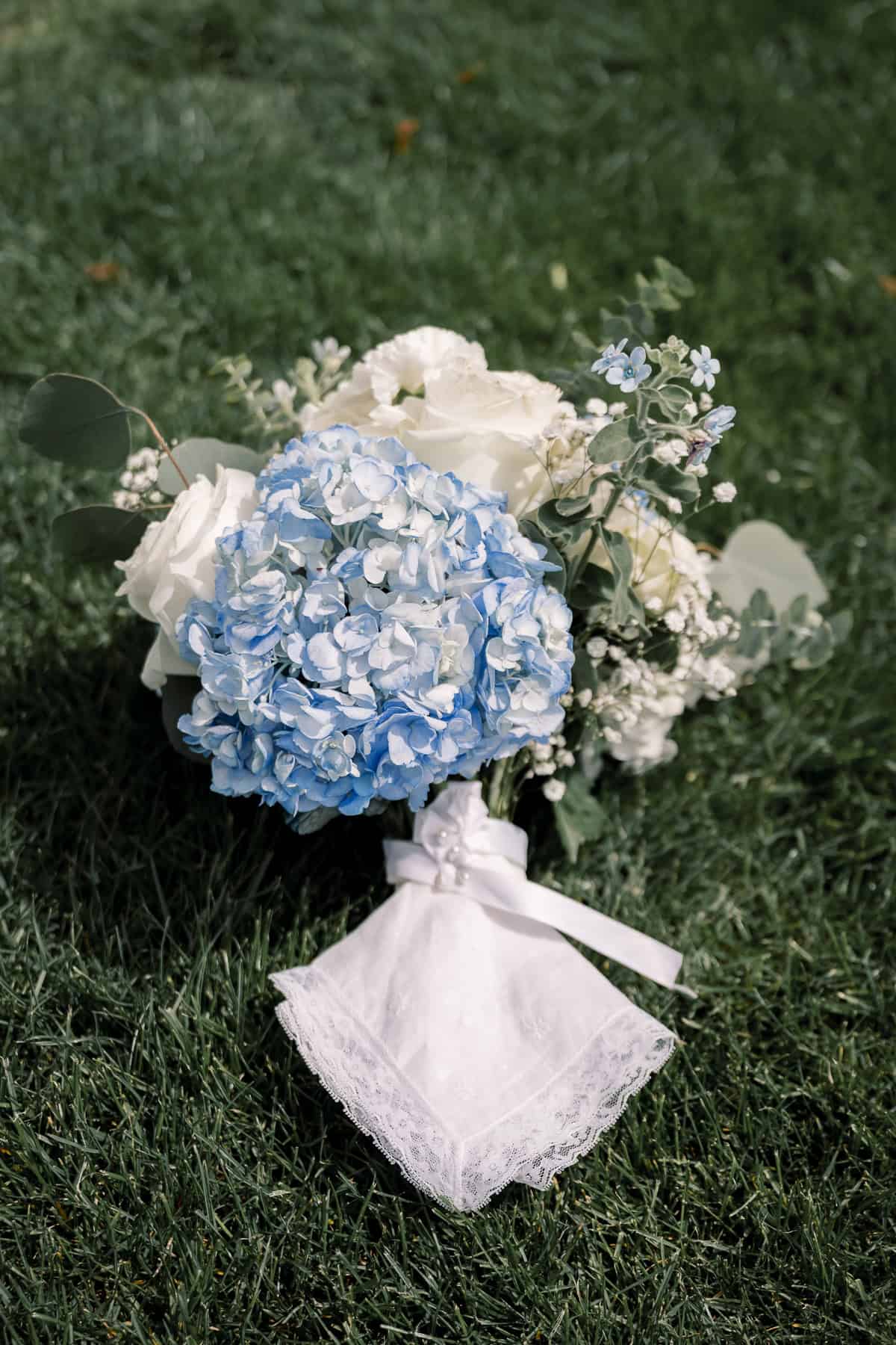 A bouquet with blue hydrangeas, white roses, greenery, and a white lace handkerchief tied with a ribbon, captured by photographer Kaity Farrell at a picturesque New England wedding, resting gracefully on the grass.