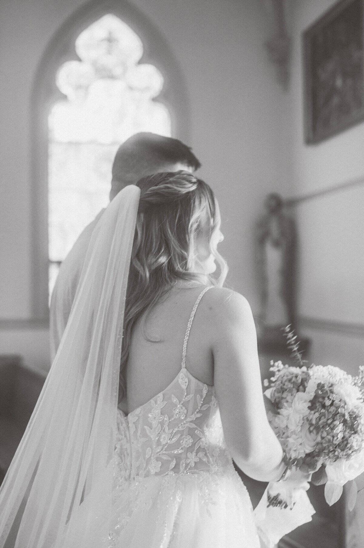 A bride in a lace dress and veil stands beside a person in formal attire, holding a bouquet, inside a church with stained glass windows, captured by photographer Kaity Farrell at this enchanting New England wedding.