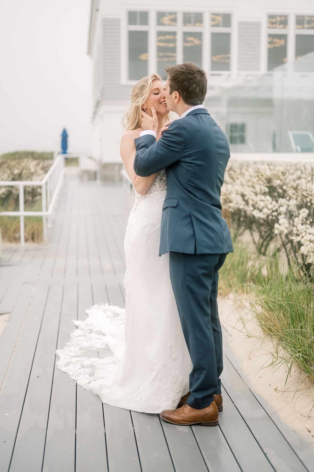 A bride and groom stand on a boardwalk at Wychmere Beach Club, smiling and holding each other, with a white building and greenery in the background—captured by wedding photographer Kaity Farrell.