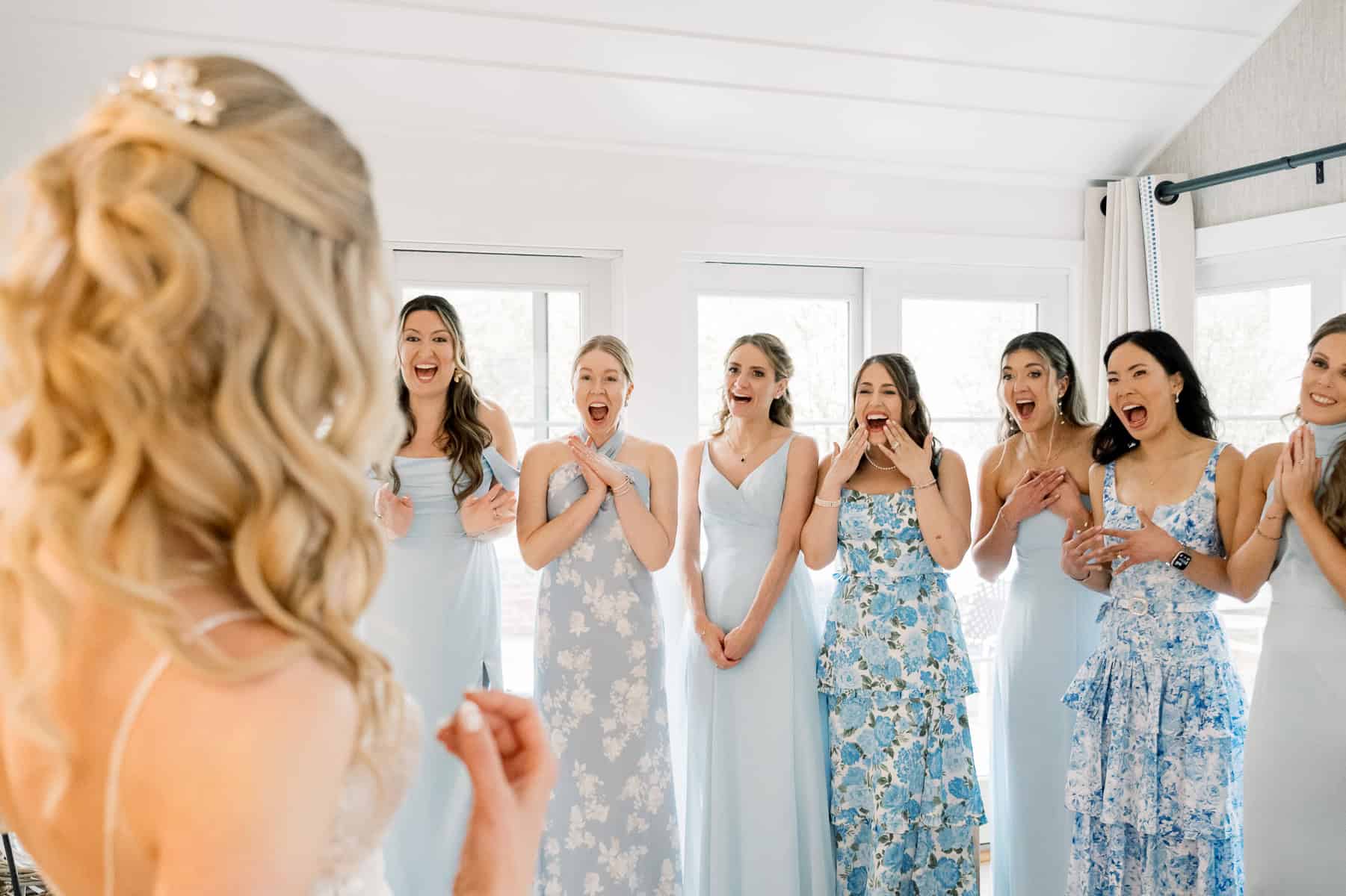 A bride faces a group of six bridesmaids in pastel blue dresses, who react with surprise and excitement in a brightly lit room, beautifully captured by Cape Cod wedding photographer Kaity Farrell.