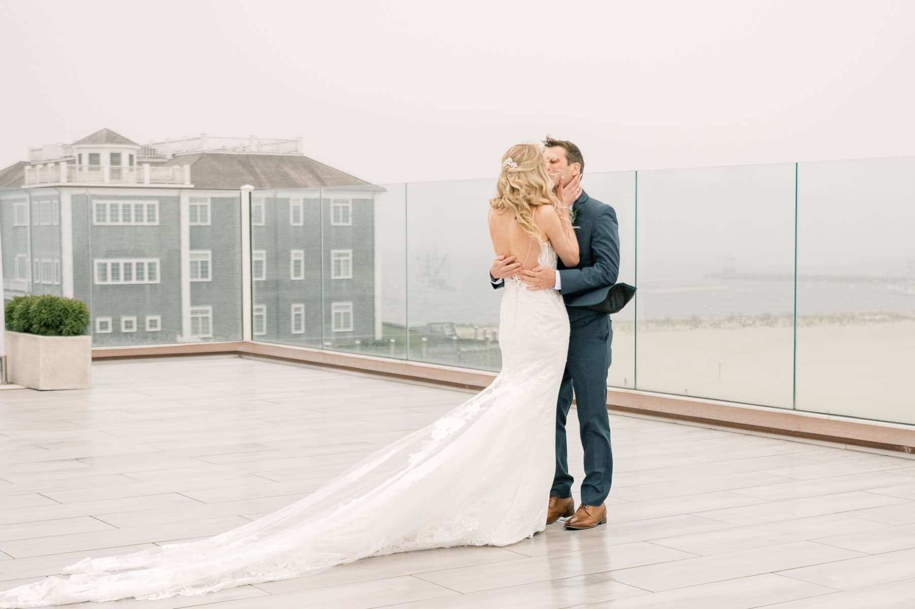 A bride in a white gown and groom in a suit embrace and kiss on a rooftop terrace with glass railings, overlooking a foggy beachfront at the Wychmere Beach Club—captured beautifully by wedding photographer Kaity Farrell.