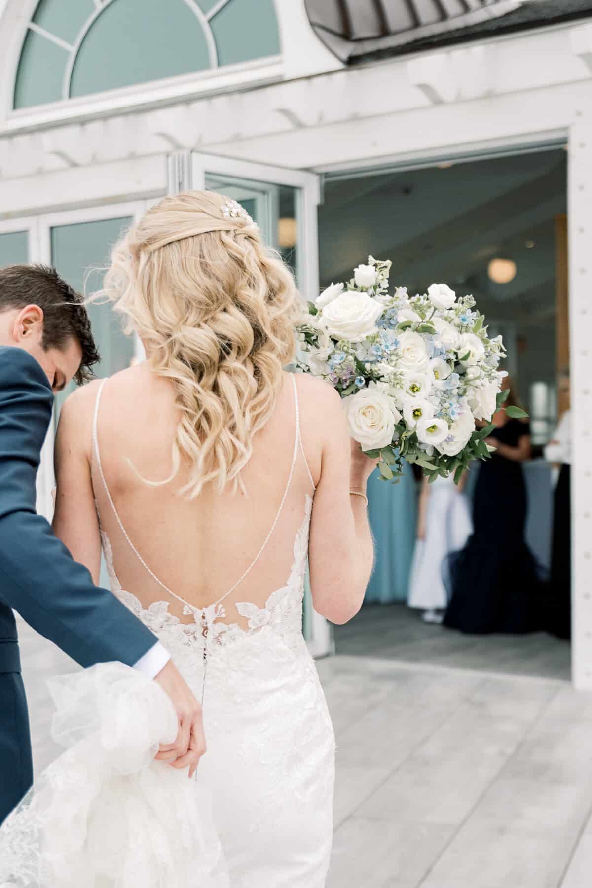 A bride in a lace wedding dress holding a bouquet of white flowers walks with a man near an open doorway at the Wychmere Beach Club, beautifully captured by Cape Cod wedding photographer Kaity Farrell.