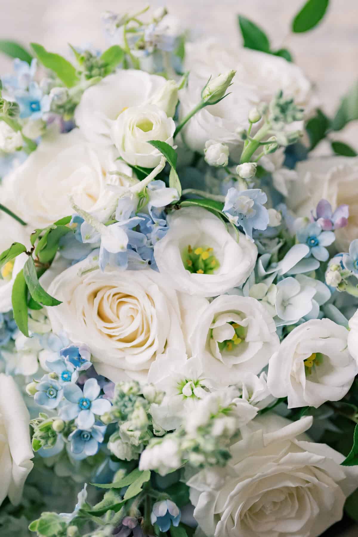 A close-up of a bridal bouquet featuring white roses, lisianthus, and light blue delphinium flowers with green foliage, beautifully captured by Cape Cod wedding photographer Kaity Farrell.