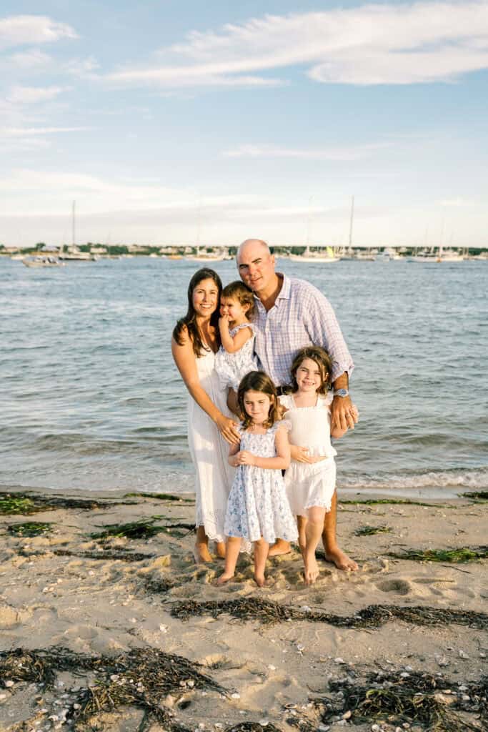 A family of five poses together on a sandy beach with the ocean and boats in the background under a partly cloudy sky during their Nantucket family portrait session with photographer Kaity Farrell.