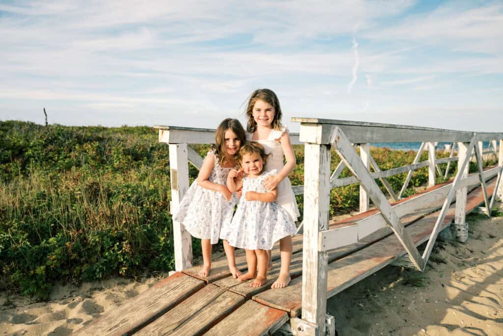 Three young girls in matching dresses stand barefoot on a wooden bridge at the beach, with greenery and blue sky in the background—capturing the charm of family portraits near Brant Point Lighthouse by photographer Kaity Farrell.