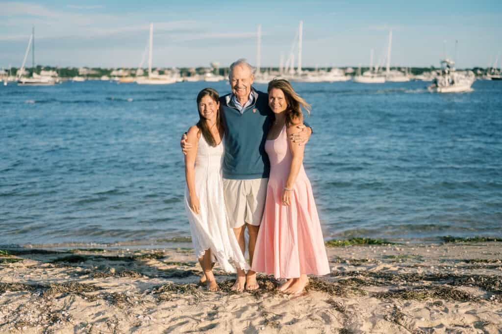 Three people stand together on a sandy beach in front of calm water and sailboats, smiling at the camera on a sunny day, perfectly captured by photographer Kaity Farrell for Nantucket lighthouse family photos.