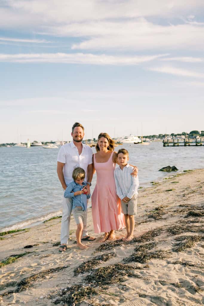 A family of four stands barefoot on a sandy Nantucket beach near the water, captured by photographer Kaity Farrell, with boats and a pier in the background under a partly cloudy sky.